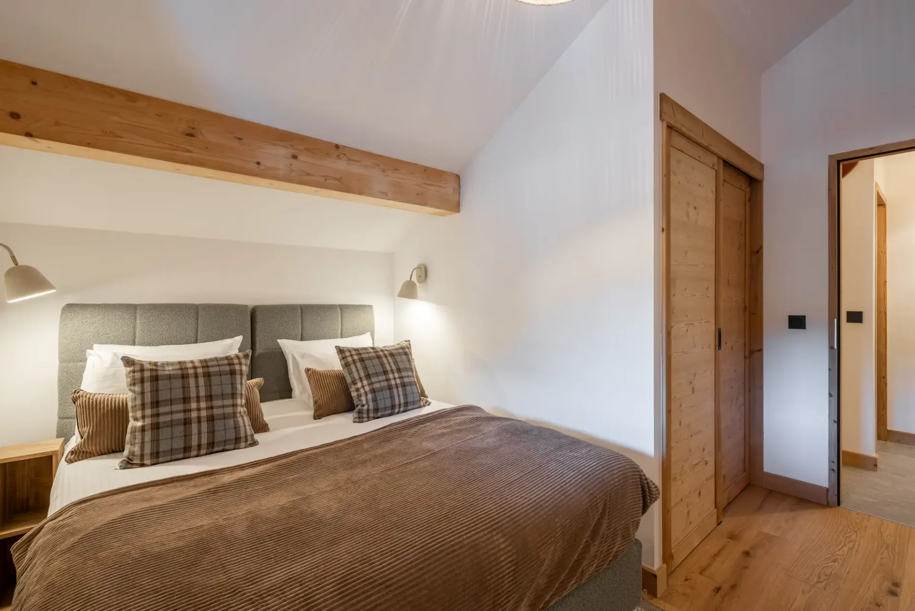 A cozy bedroom with a double bed, plaid pillows, a wooden beam ceiling, a window with a view, and a dresser with a lamp.