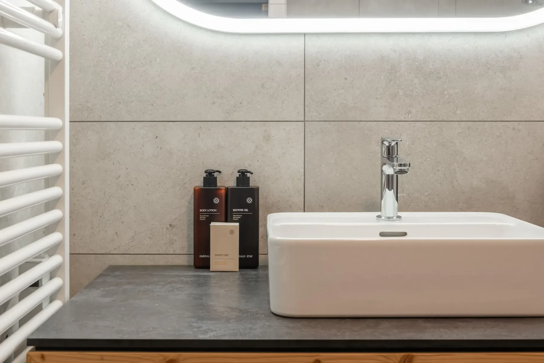 Modern bathroom with gray tile walls, wall-mounted toilet, glass-enclosed bathtub and shower, sink on a wooden vanity, and open shelving.