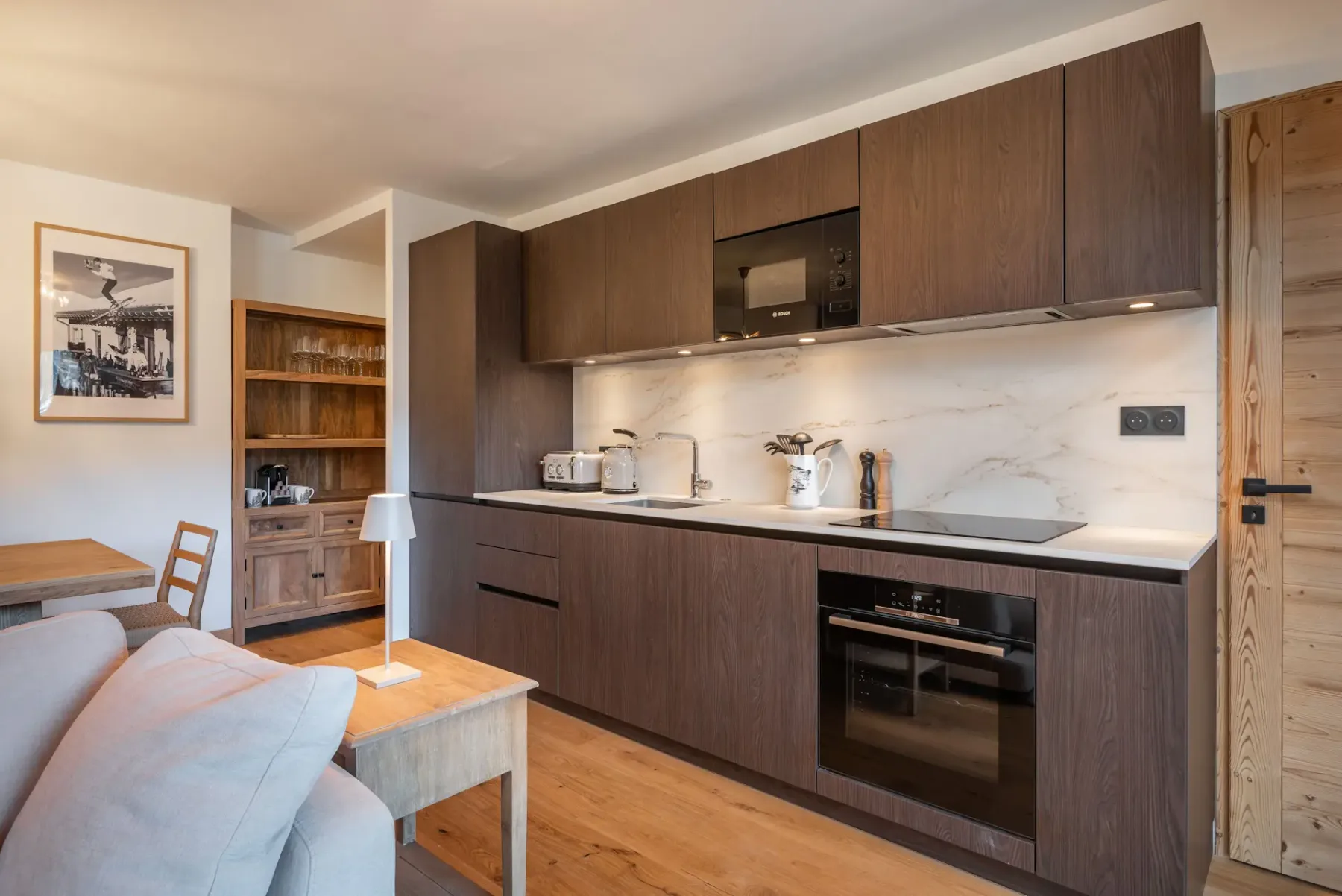 Modern dining area with a wooden table and six chairs, minimalist decor, flower centerpiece, wall art, and open kitchen with dark cabinets in the background.