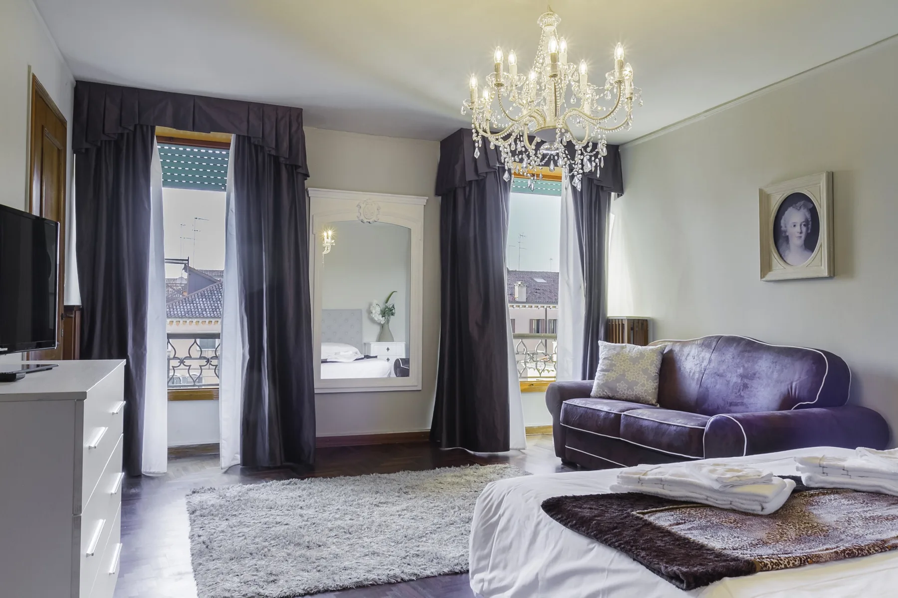 A bedroom with a bed, purple seating, and tall curtained windows.