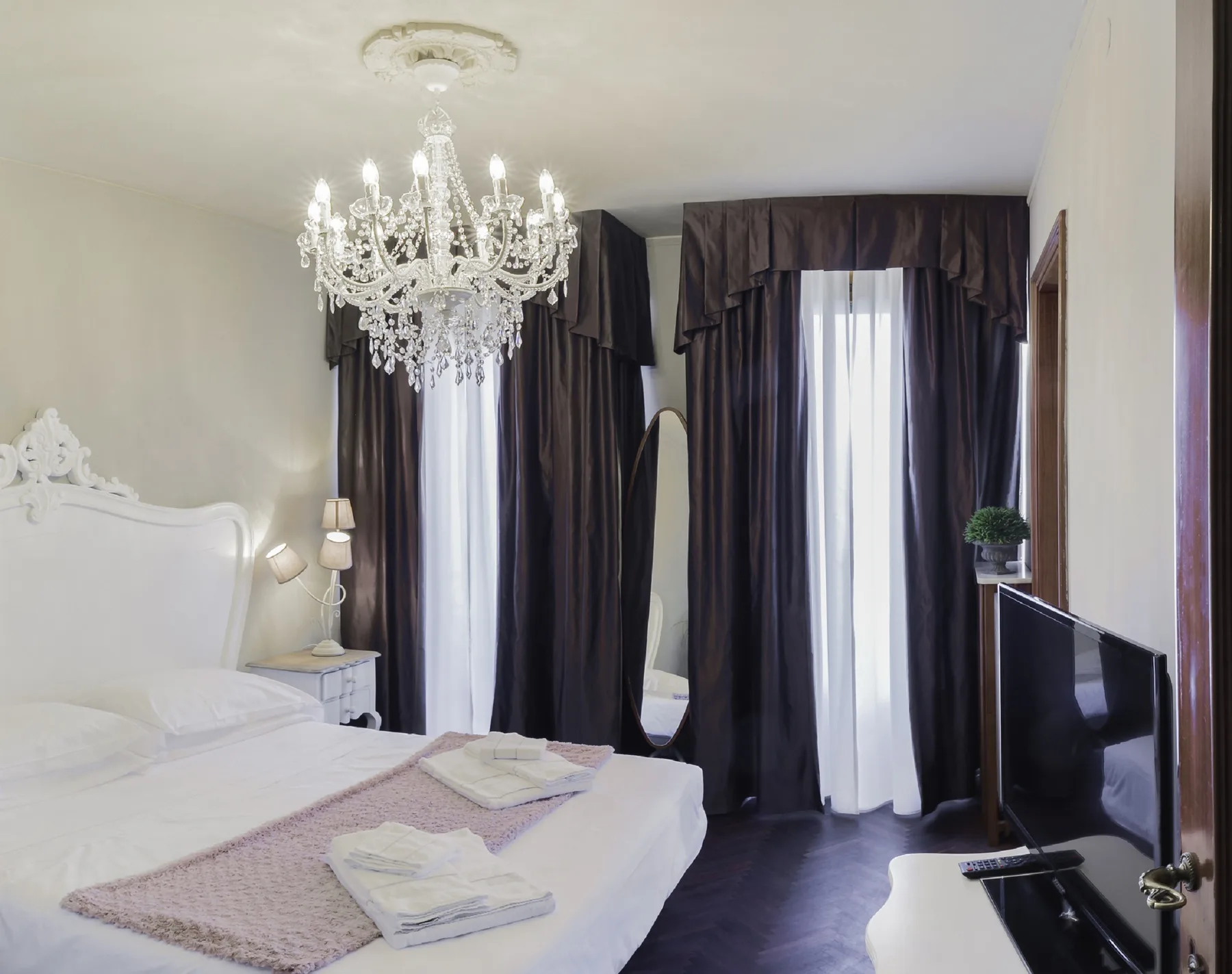 A bedroom with a chandelier, a bed, and dark curtains framing tall windows.
