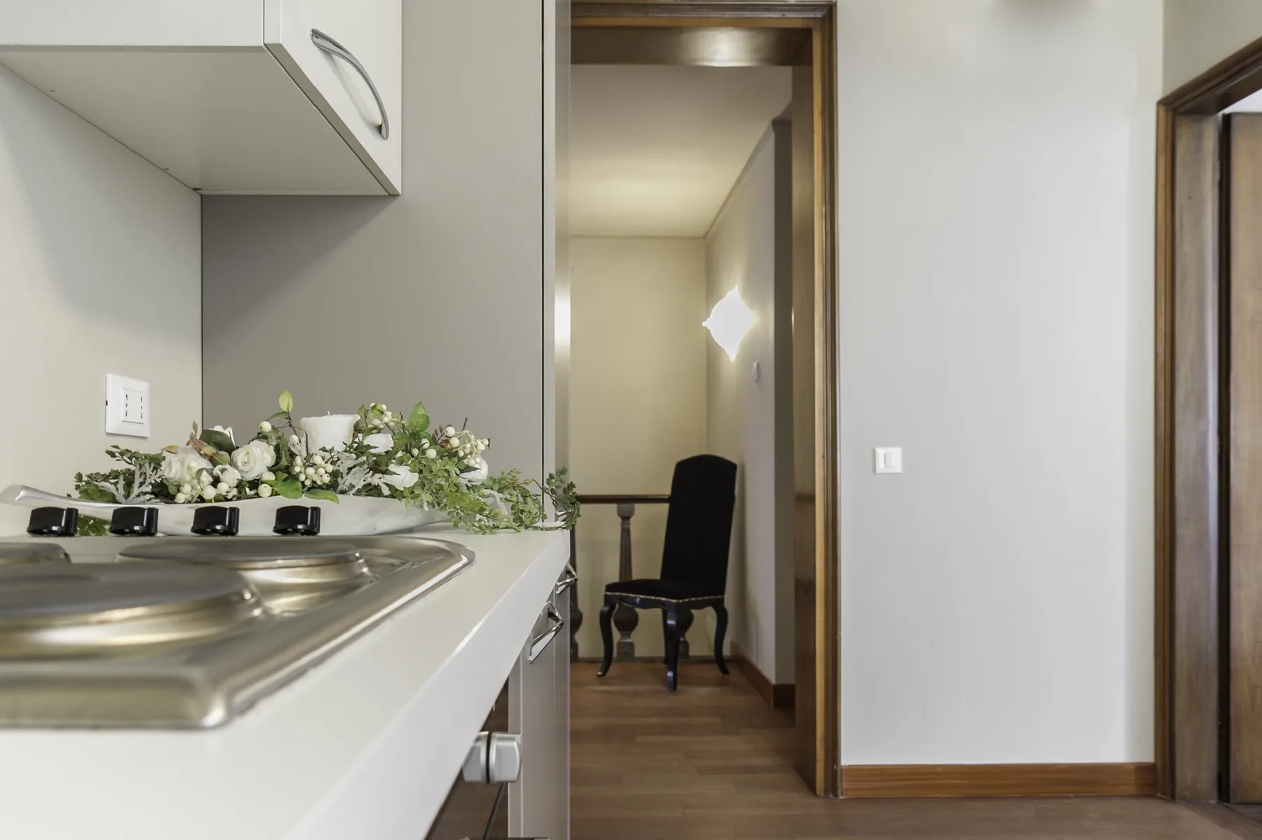 A kitchenette opens onto a hallway with a chair and wall light.