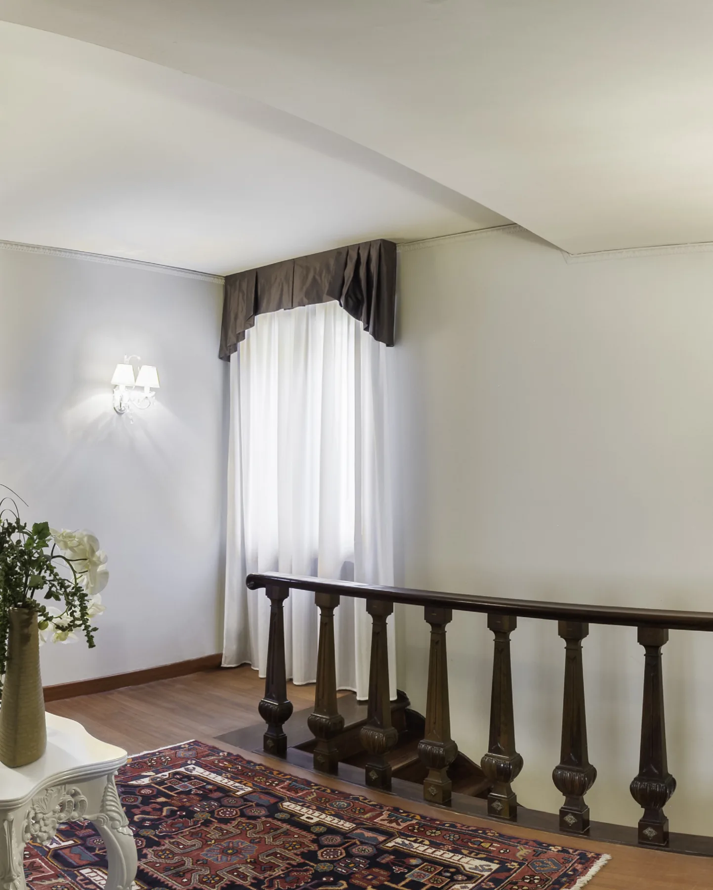 A stair landing with a dark wooden railing, patterned rug, and curtained window.