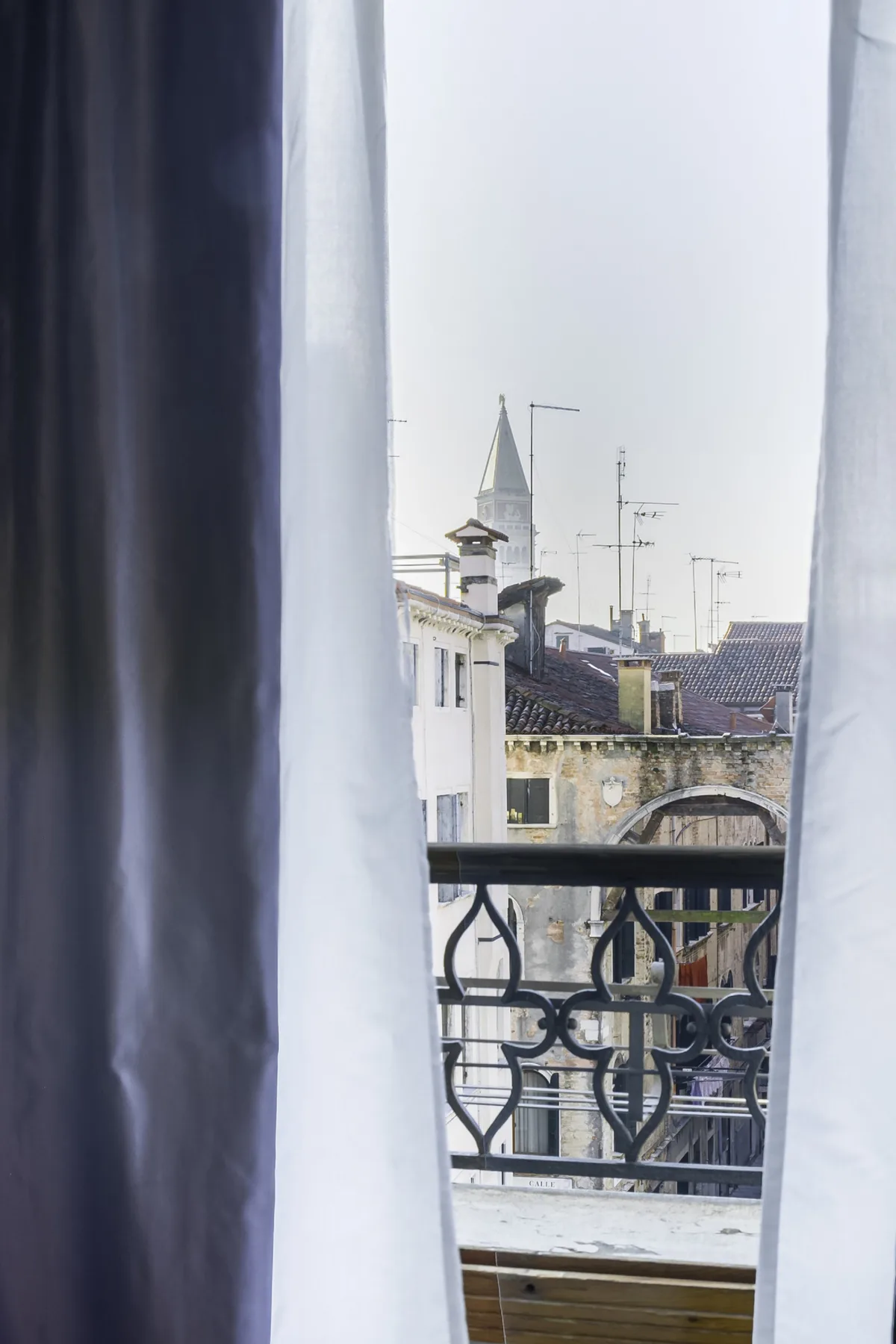 A city view through open curtains, with rooftops and a balcony railing.