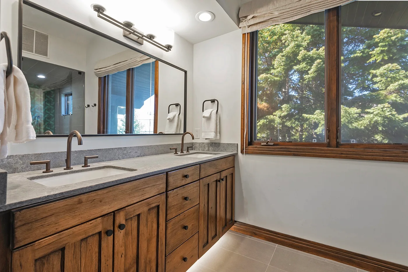A bathroom vanity with two sinks sits beside a large window overlooking leafy trees.