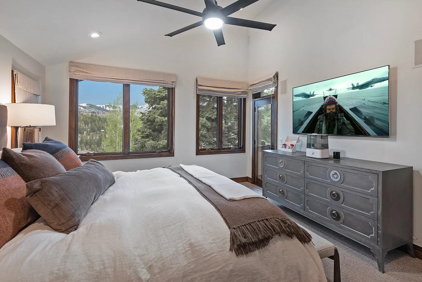 A bedroom with a neatly made bed, large windows, and a dresser with a television.