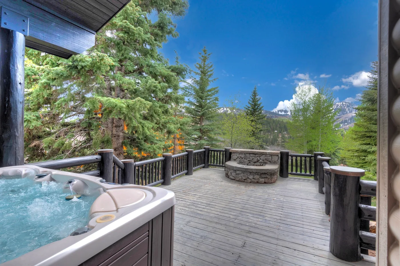 A wooden outdoor deck with a hot tub and round stone fire pit overlooks trees.