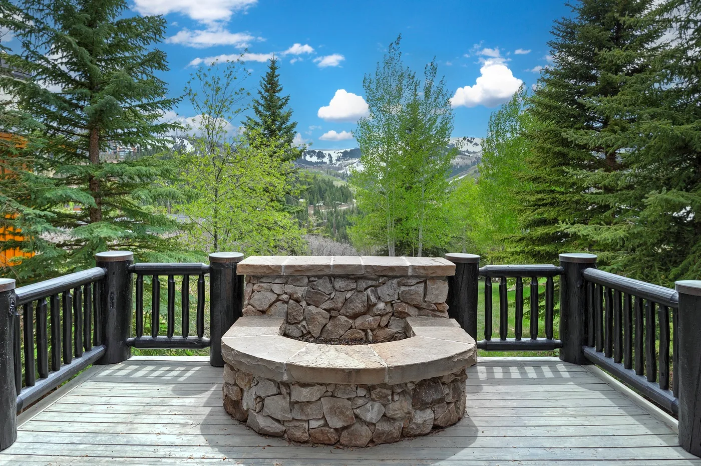 An outdoor deck overlooks a green forest, with a stone fire pit in the foreground.