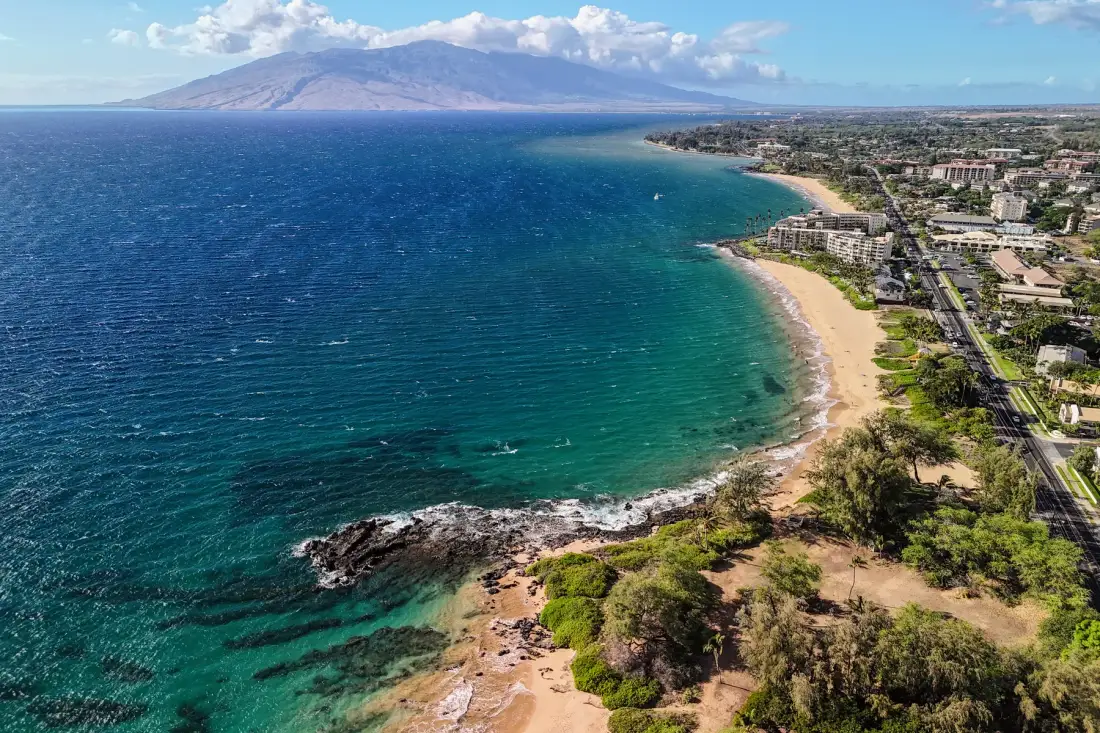 Aerial view of a curving coastline, with blue water, a sandy beach, and nearby buildings.