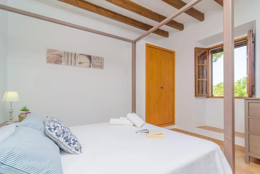 A bright bedroom with a bed, wooden ceiling beams, and an open window.
