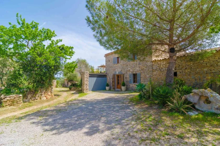 A stone house stands beside a gravel driveway beneath large trees.