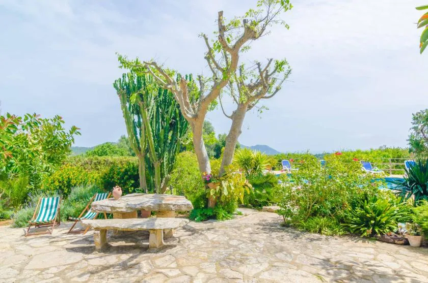 A stone patio with picnic tables sits among trees and lush garden plants.