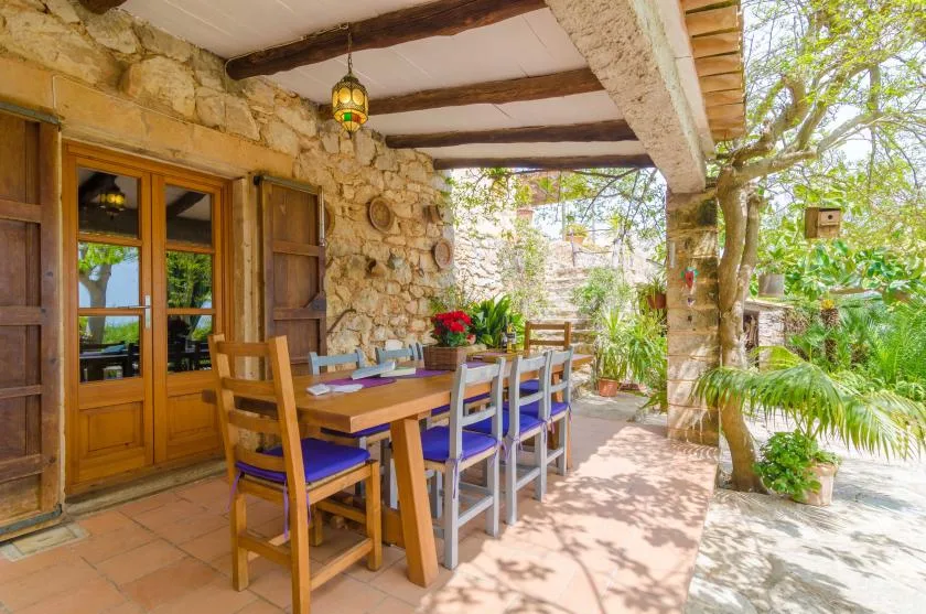 A covered outdoor dining area has a wooden table and chairs beside a stone wall.
