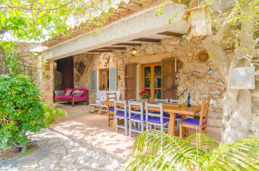 A covered outdoor dining area with a long table and colorful chairs beside a stone wall.