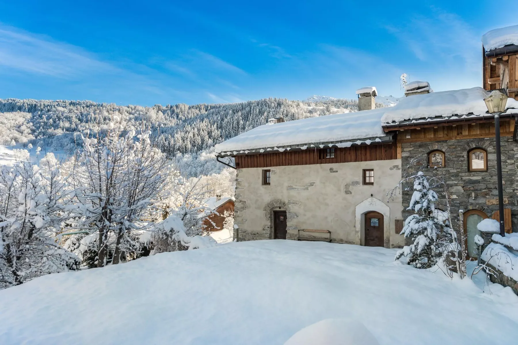 A snow-covered chalet stands beside snowy trees against a forested mountain.