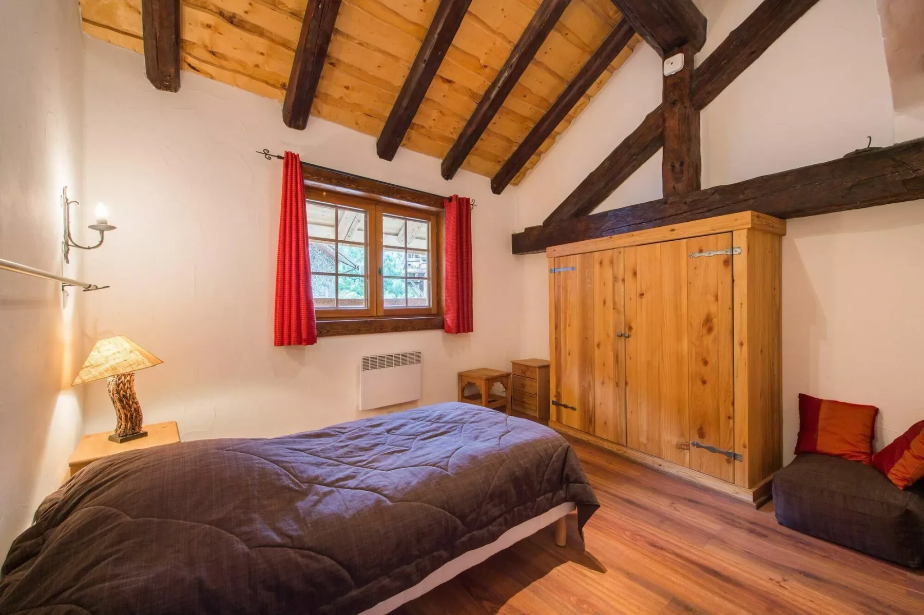 A bedroom with a bed, wooden wardrobe, and a window framed by red curtains.
