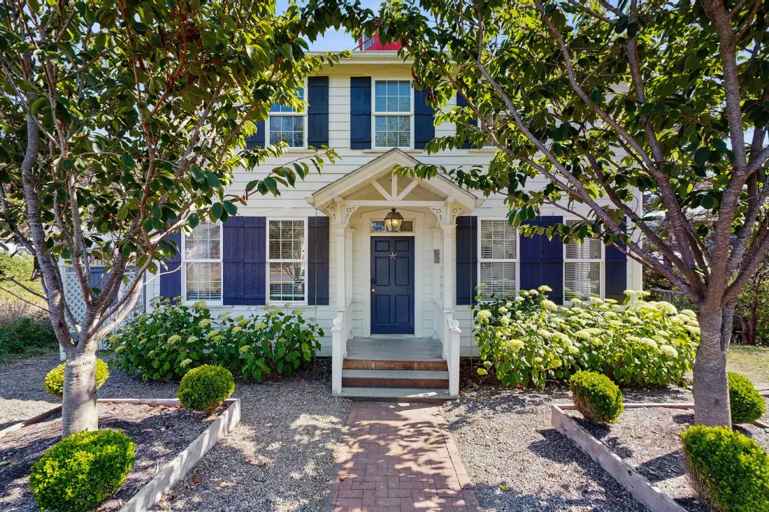 A two-story house entrance with a brick walkway and leafy trees.