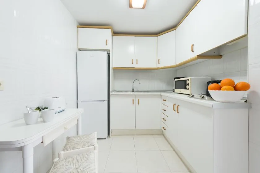 A bright white kitchen with cabinets, a refrigerator, and a bowl of oranges.