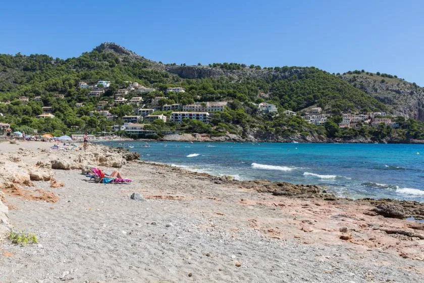 A sandy beach meets clear blue water below a green hillside.