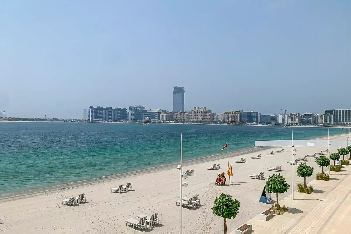 A sandy beach borders turquoise water, with lounge chairs and buildings across the bay.