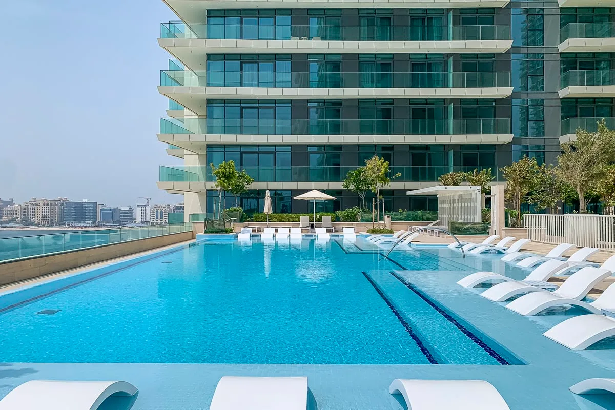A rooftop swimming pool beside a modern glass building, with lounge chairs along the water.