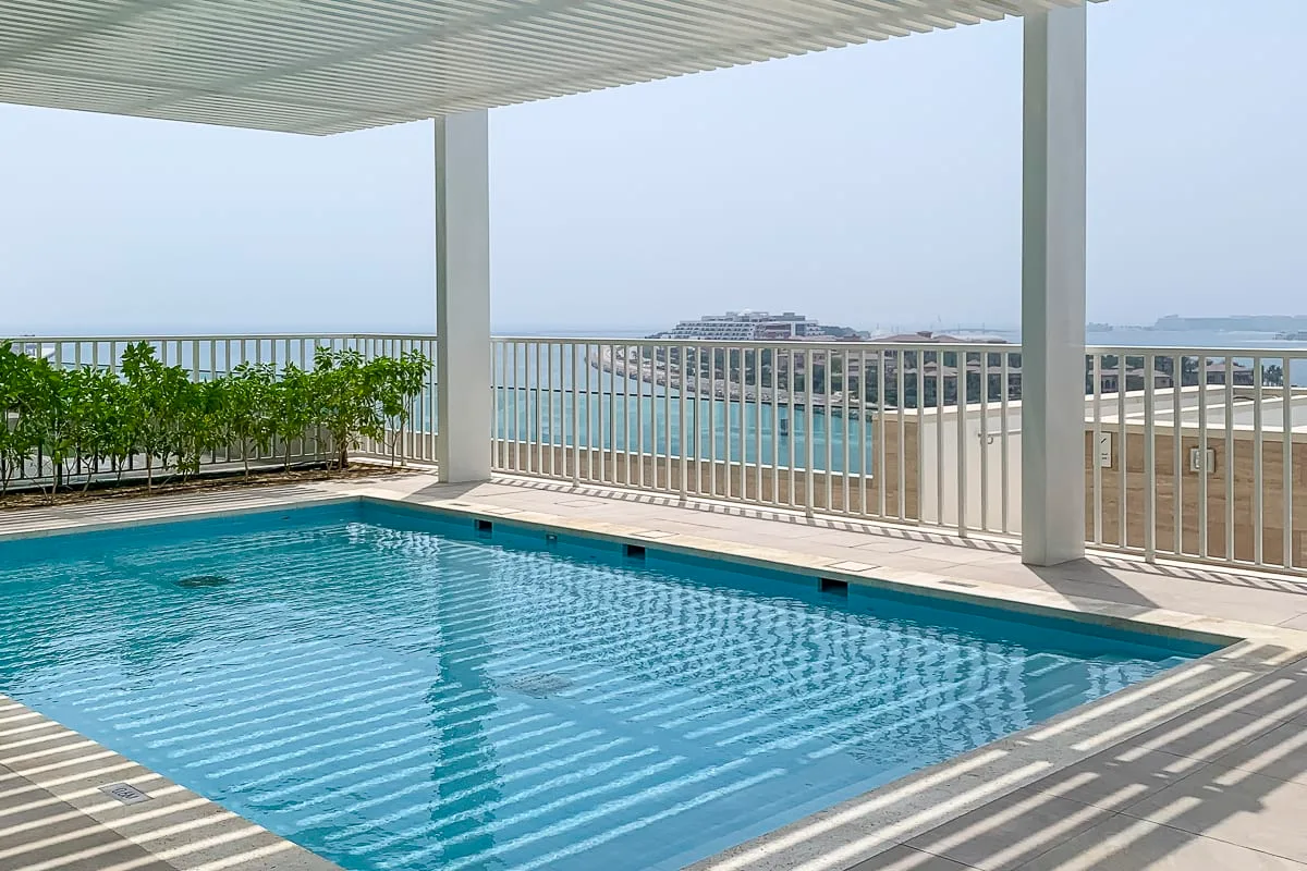 A swimming pool sits beneath a shaded terrace beside a railing overlooking the water.