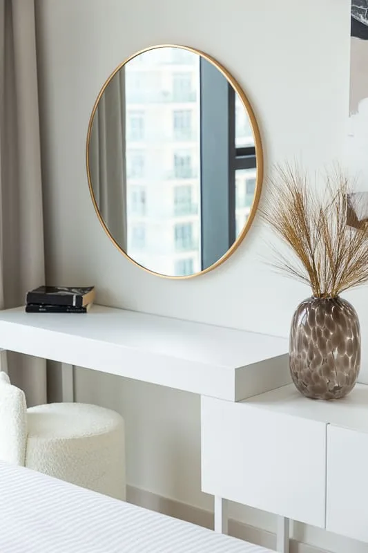 A bright bedroom corner with a white desk, round mirror, and decorative vase.