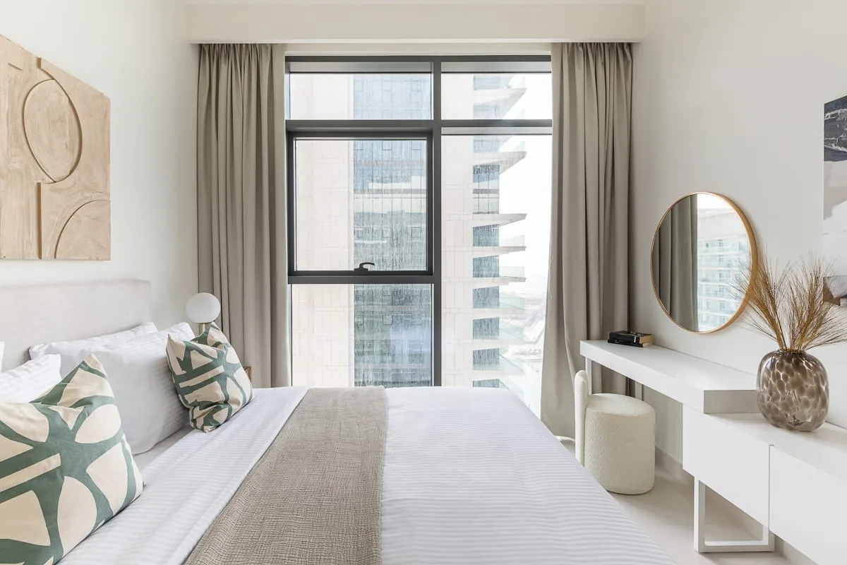 A bright bedroom with a large window, a bed, and a dressing table.