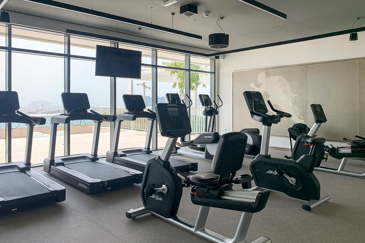 A gym with treadmills and stationary exercise bikes beside large windows.