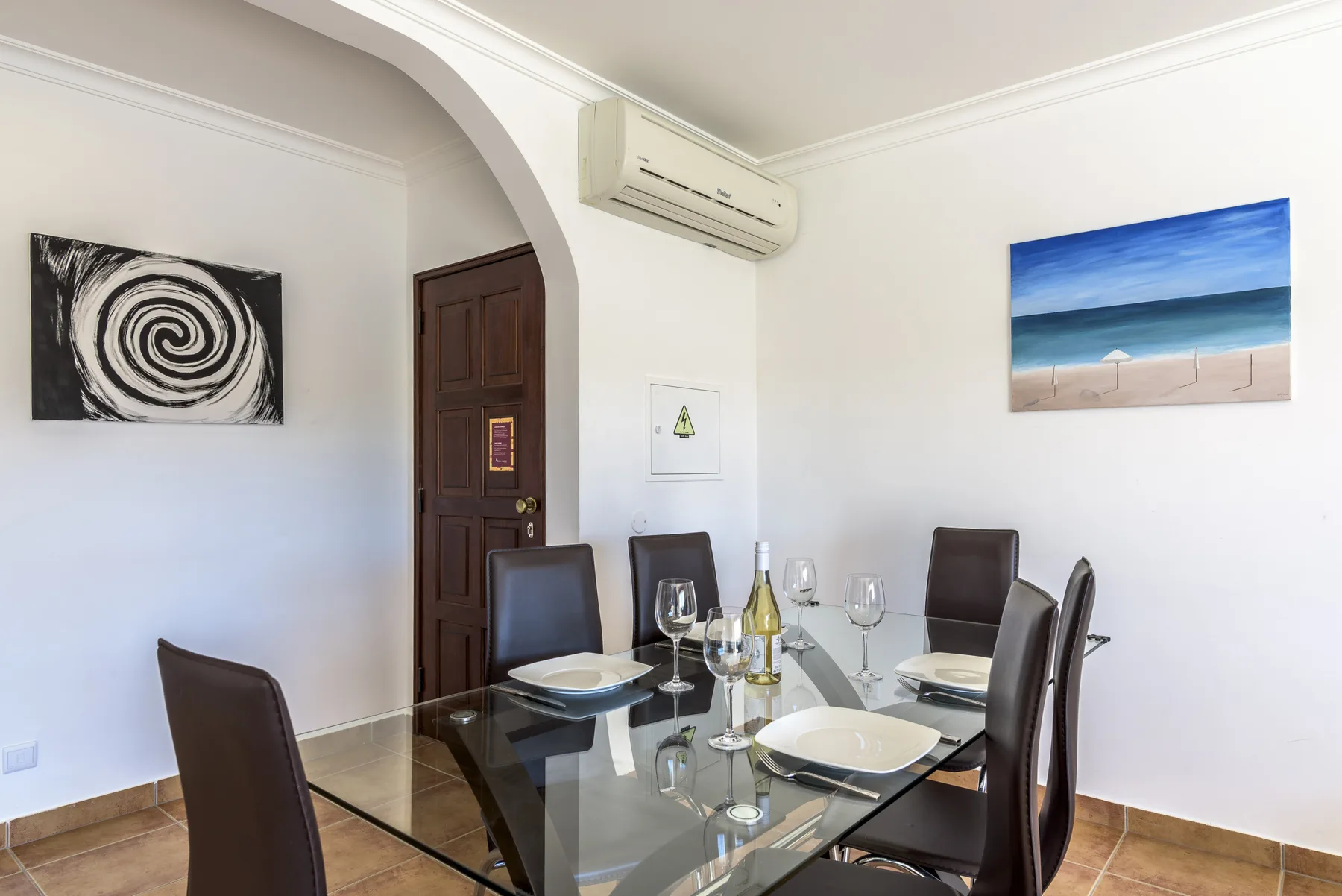 Comedor con mesa de cristal y asientos oscuros bajo un aire acondicionado en la pared.