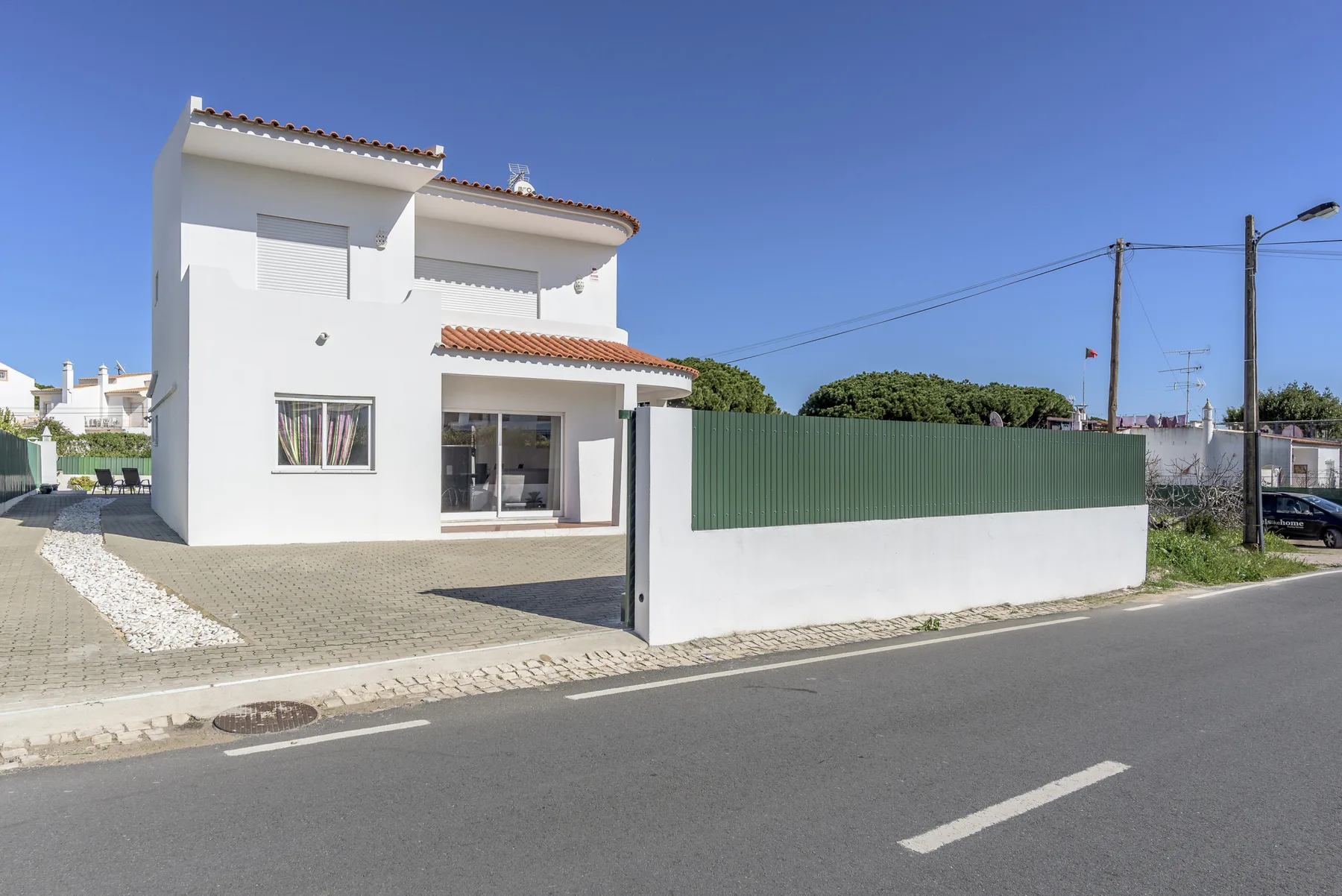 Una casa blanca de dos plantas se encuentra detrás de una valla verde junto a la carretera.