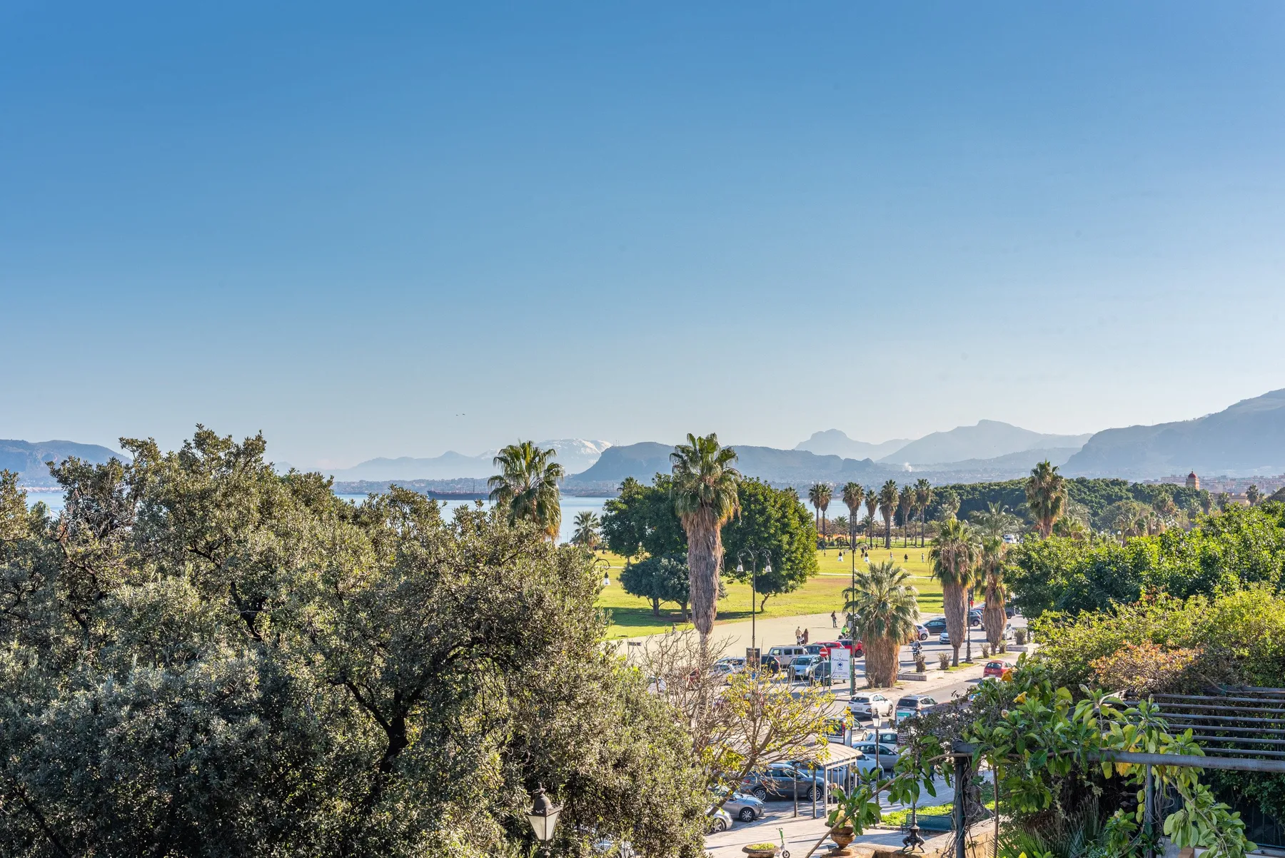 A sunny landscape overlooks a lake, with trees, palms, and mountains in the distance.