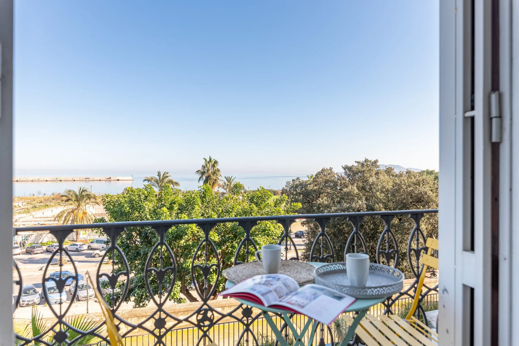 A balcony overlooks leafy trees, palm trees, and a distant coastline.