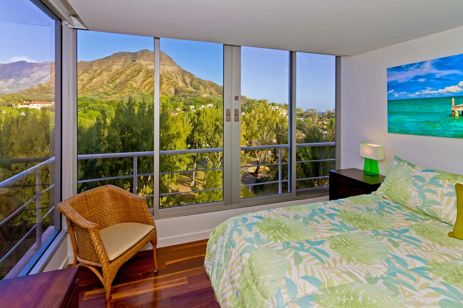 A bedroom with a bed, a wicker chair, and large windows overlooking greenery and mountains.