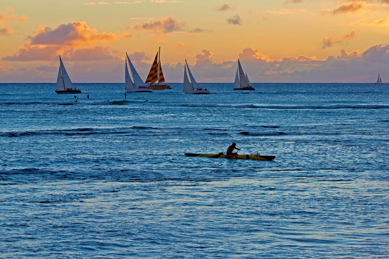 Veleros cruzan el mar al atardecer mientras una persona rema en kayak en primer plano.