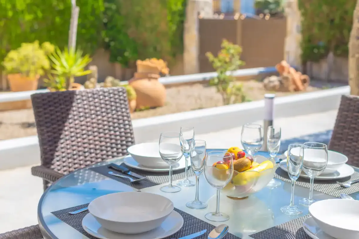 An outdoor dining table is set with plates and glasses beside a garden.