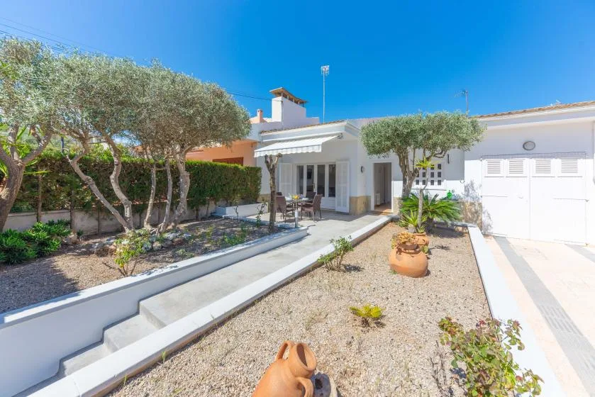 A white house opens onto a gravel garden with trees and a small patio.