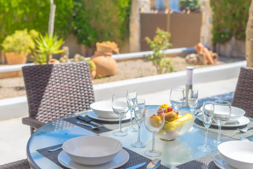 An outdoor dining table is set with plates, glasses, and a fruit bowl.