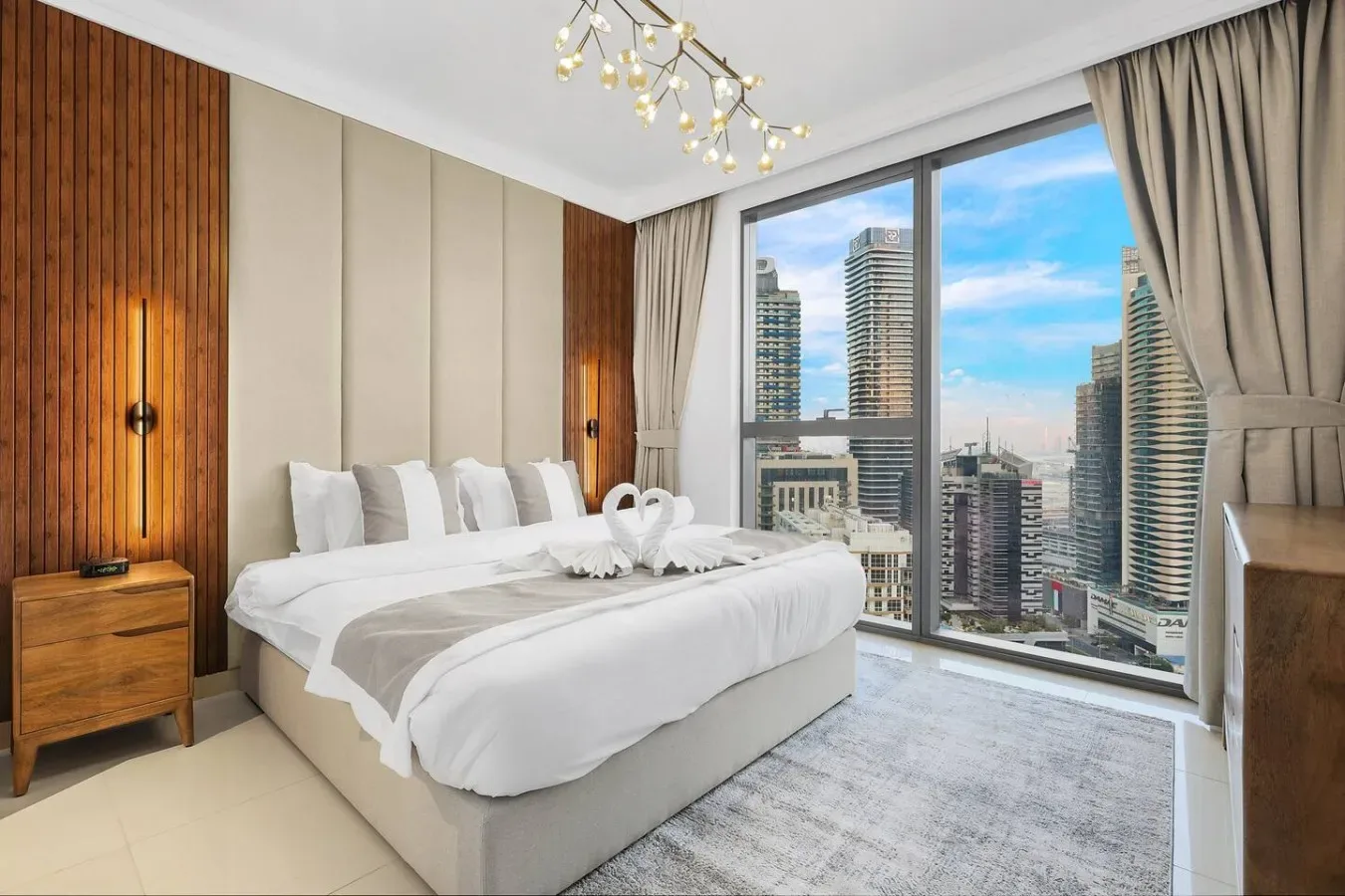 Un dormitorio luminoso con una cama bien tendida junto a grandes ventanales con vistas a edificios de la ciudad.