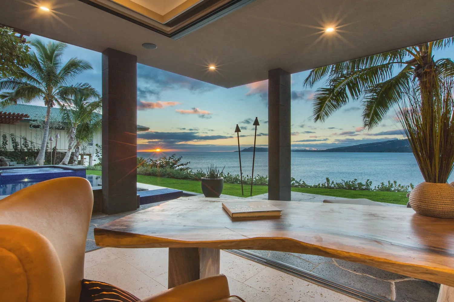 A covered patio overlooks a pool, palm trees, and the water at sunset.