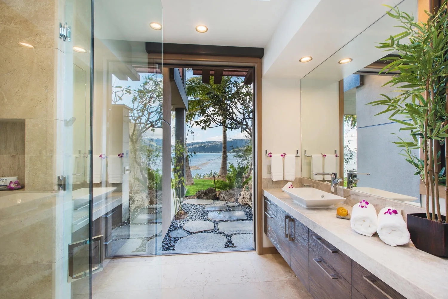 A bright bathroom with a long vanity and a glass shower opening onto a garden view.