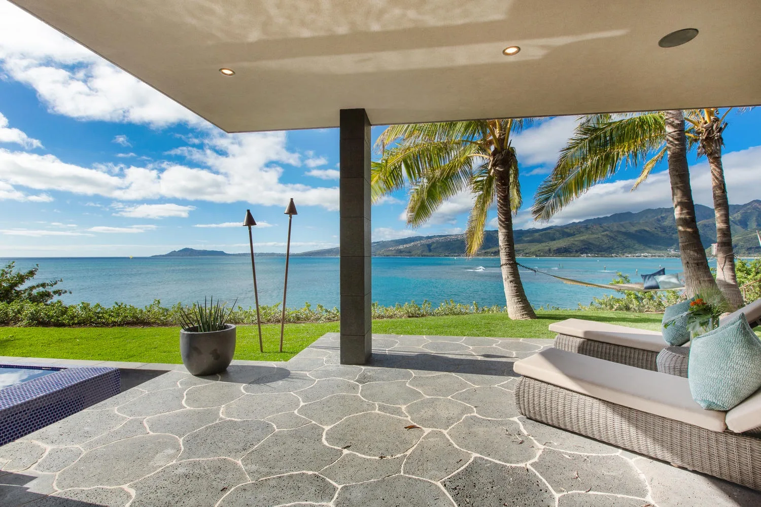 A covered patio with seating overlooks the ocean and palm trees.