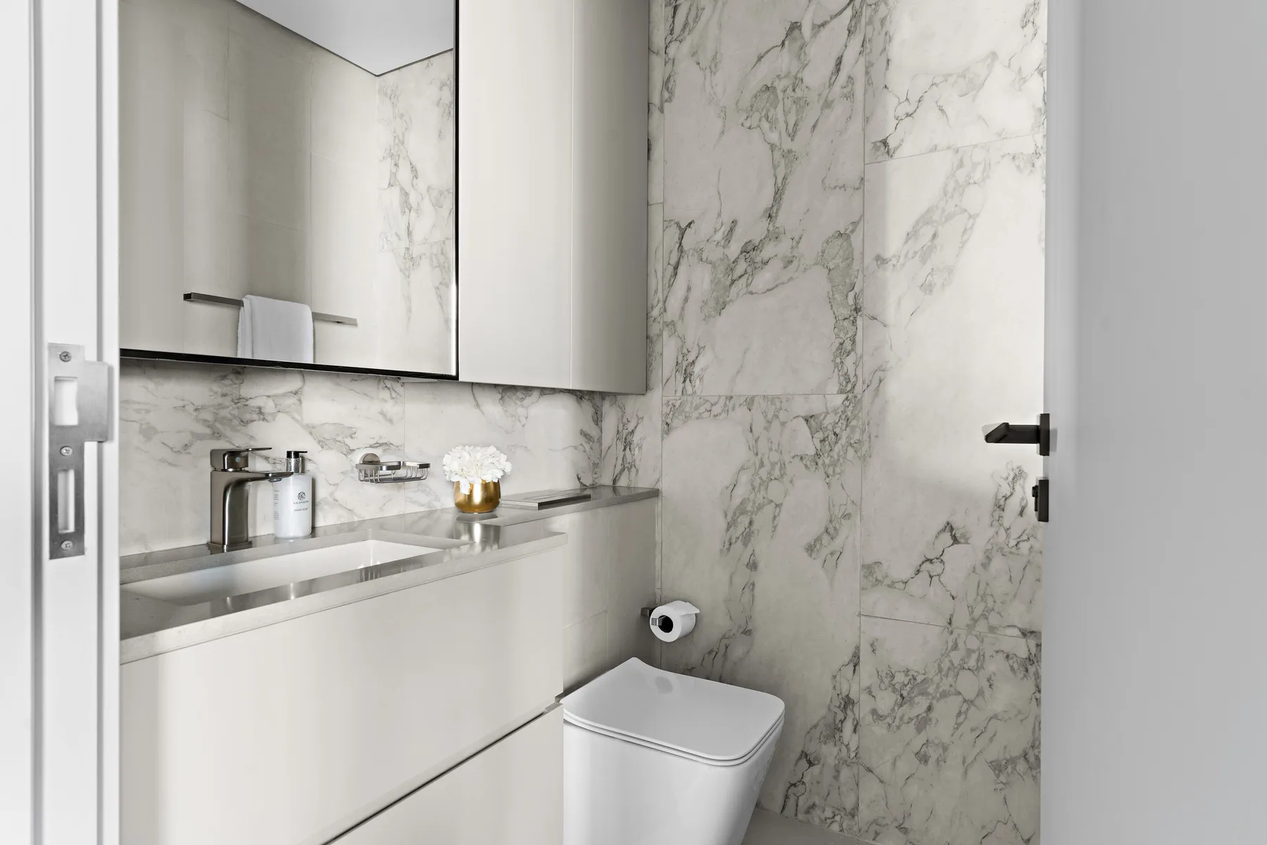 A bathroom with a sink vanity, mirror, toilet, and marble-patterned walls.