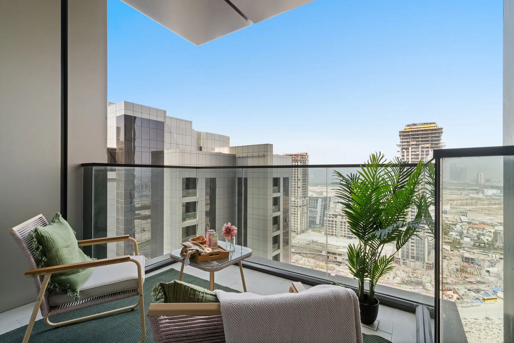 A balcony seating area with a glass railing and potted palm overlooks nearby buildings.