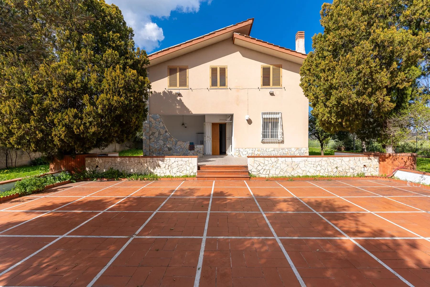 Una casa está detrás de una cancha exterior embaldosada, con árboles a ambos lados.