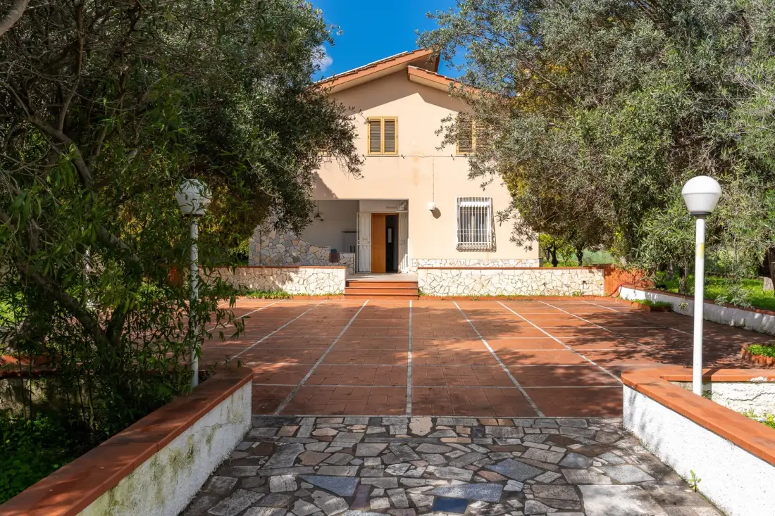 Una casa se alza al fondo de un patio pavimentado, rodeada de árboles.