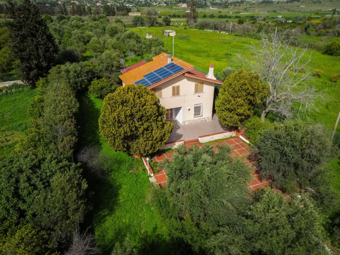 Vista aérea de una casa con paneles solares en el tejado, rodeada de campos verdes y árboles.