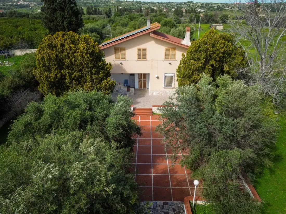Una casa se encuentra al final de un camino de baldosas rojas, rodeada de árboles y vegetación.