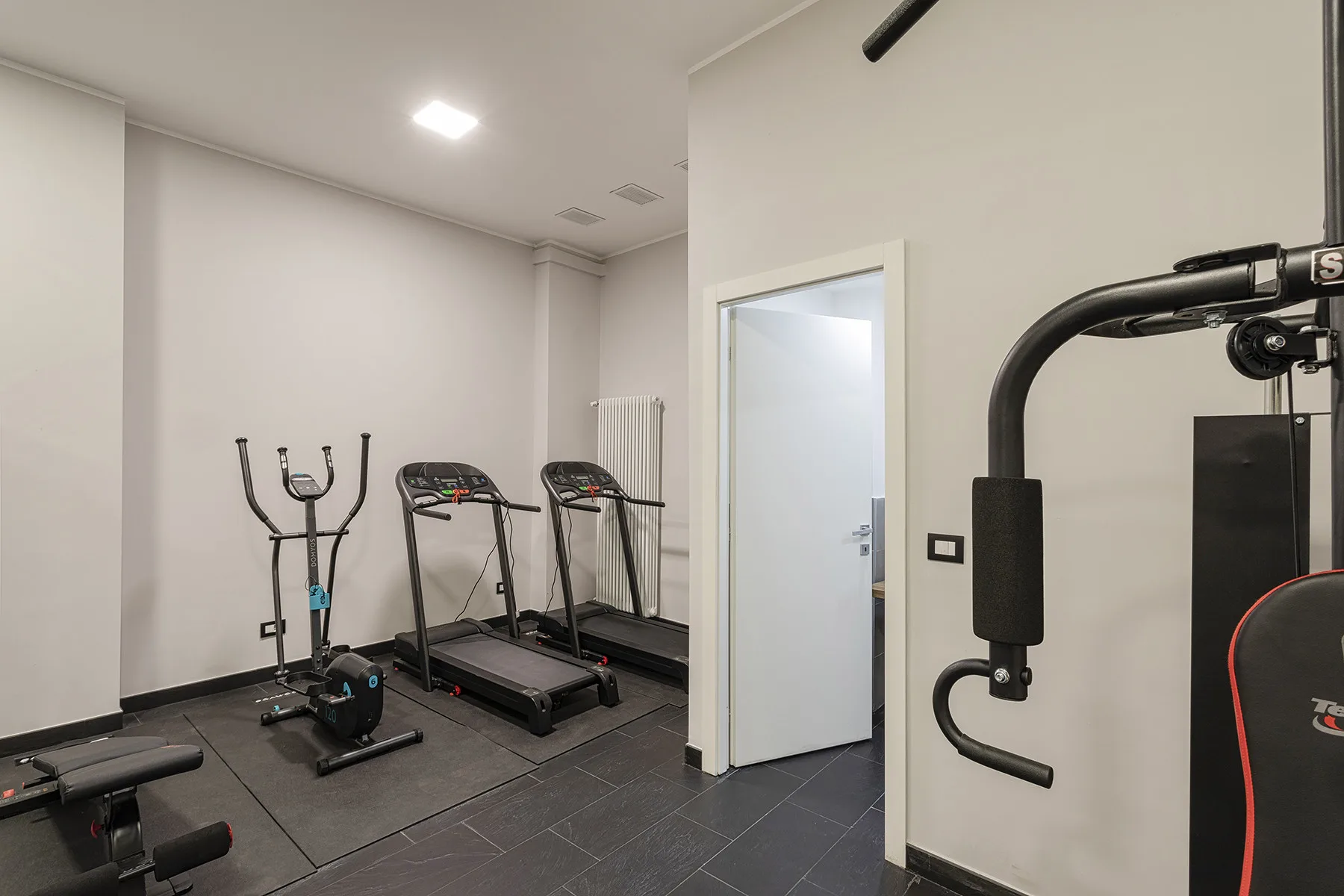 Un pequeño gimnasio con cintas de correr y equipo de ejercicio sobre un suelo de baldosas oscuras.