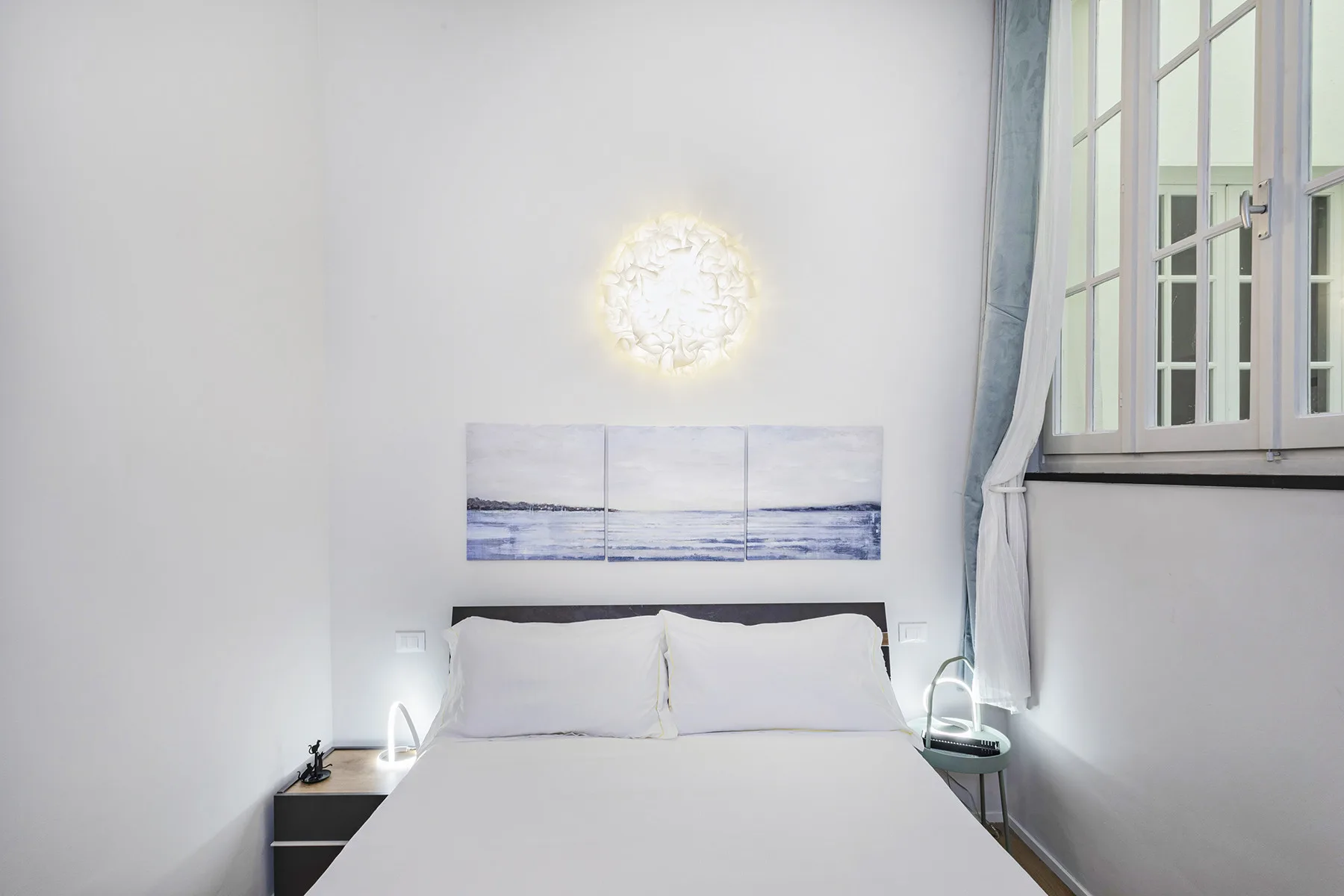 Un dormitorio luminoso con una cama bajo un cuadro marino de tres paneles.